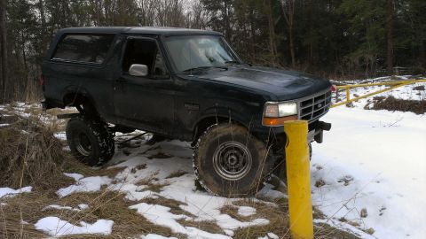 1995 Ford Bronco - A dark green Ford Bronco navigates a snowy, wooded area with a yellow gate in the background.