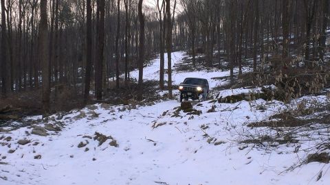 1995 Ford Bronco - A dark SUV navigates a snowy path through a dense forest with bare trees.