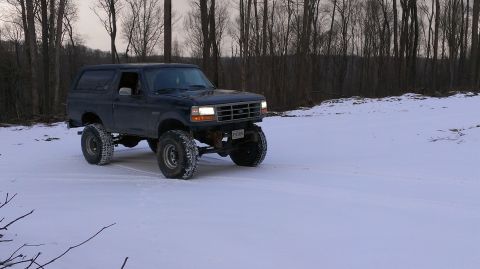1995 Ford Bronco - A dark SUV with a lifted suspension drives through a snowy landscape with trees in the background.