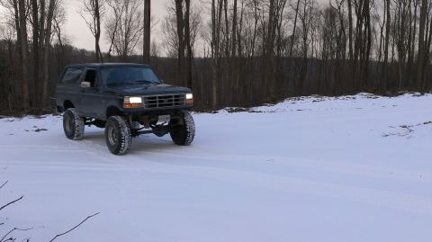 1995 Ford Bronco - Lifted 1995 Ford Bronco navigates a snow-covered landscape with a wooded area behind it.
