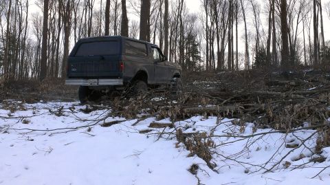 1995 Ford Bronco - A Ford Bronco navigates a snowy, wooded terrain with fallen trees and branches scattered across the ground.
