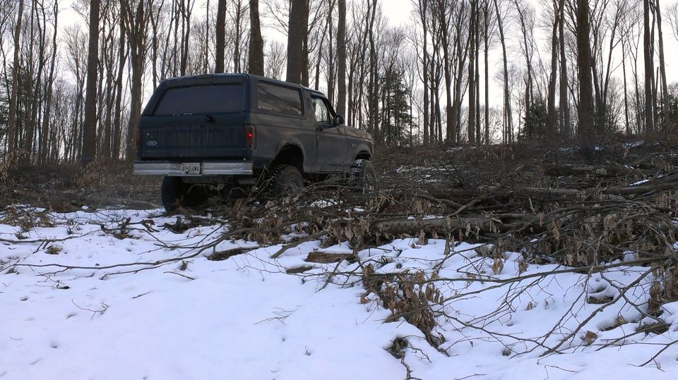 Dark-colored Ford Bronco driving through snow and fallen branches in a wooded area