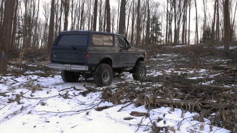 1995 Ford Bronco - A dark blue SUV navigates a snowy, debris-strewn forest path.