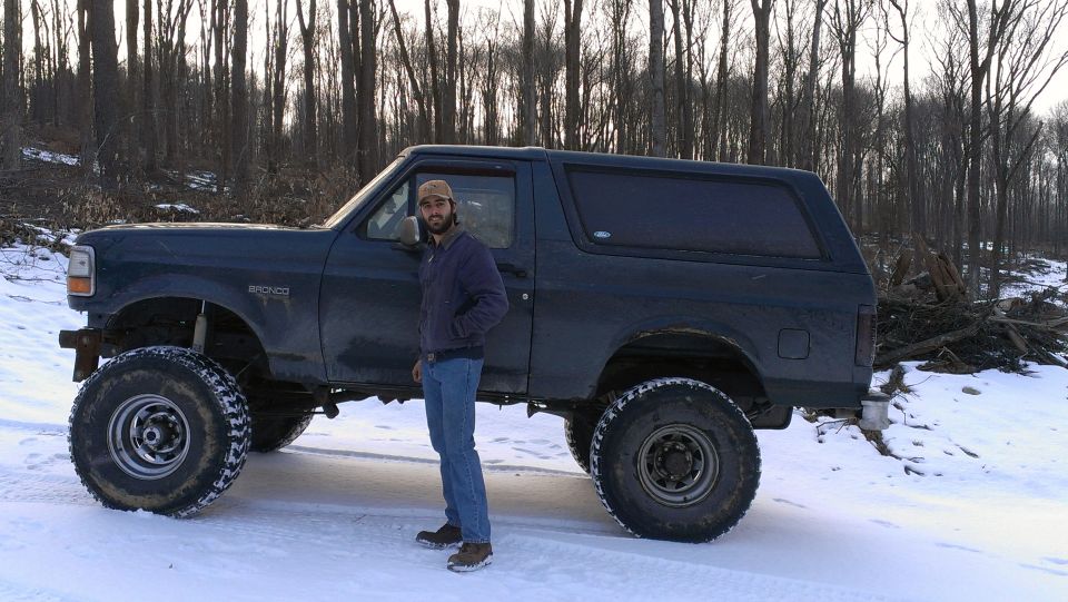 Dark blue Ford Bronco with large tires and a man standing next to it in a snowy wooded area