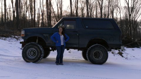 1995 Ford Bronco - A dark 1995 Ford Bronco with oversized tires is parked in a snowy, wooded area with a woman standing beside it.