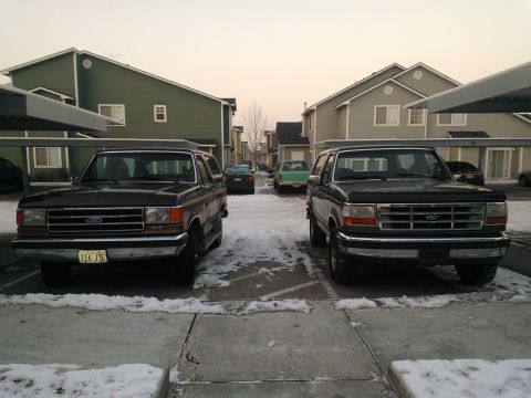 1992 Ford Bronco - Two Ford Broncos are parked in a lot with snow on the ground, with apartment buildings in the background.