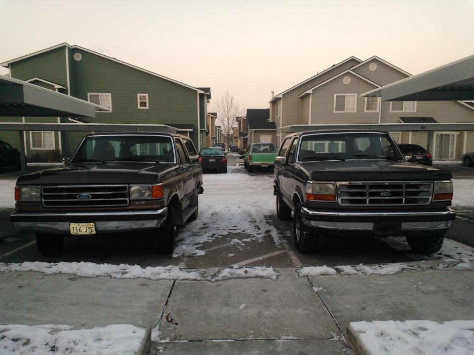 Two Ford Broncos parked in front of apartment buildings. One is dark, the other is dark gray. A green car is in the background