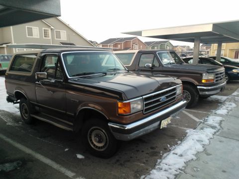 1992 Ford Bronco - Two trucks, a Ford Bronco and a Ford F-Series, are parked side-by-side in a lot with snow on the ground.