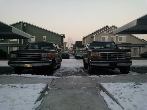 1992 Ford Bronco - Two 1992 Ford Broncos are parked front-facing in a snowy lot with apartment buildings in the background.