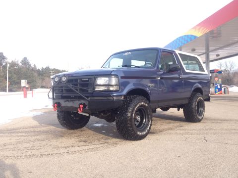 1996 Ford Bronco - A blue 1996 Ford Bronco with a lifted suspension and custom front bumper is parked outside a gas station in snowy conditions.