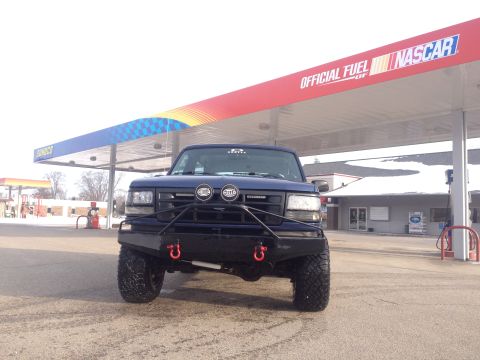 1996 Ford Bronco - A blue Ford truck with a heavy-duty bumper and large tires is parked at a gas station under a NASCAR-branded awning.