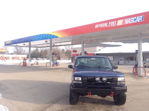 1996 Ford Bronco - Blue Ford F-Series truck with aftermarket bumper and lights at a gas station.