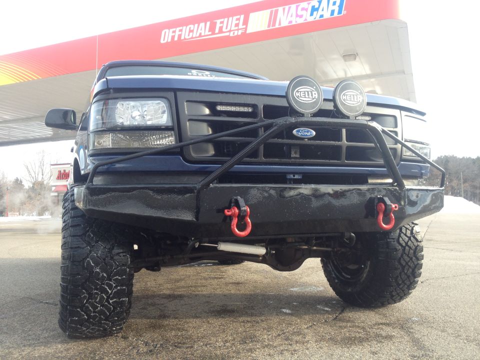Front view of a blue Ford F-Series truck with a custom bumper and Hella lights. NASCAR branding visible in background