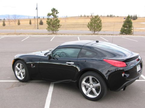 1995 Ford Bronco - Black Pontiac Solstice GXP Coupe parked in a lot with trees and hills in the background.