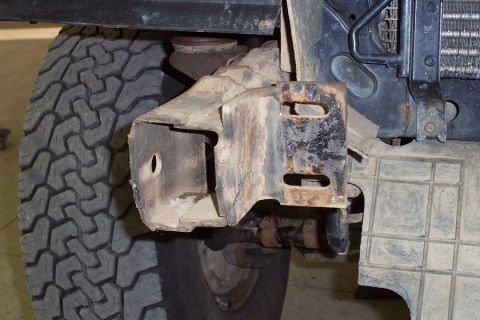 1996 Ford Bronco - Close-up of a rusty steel front suspension/frame bracket on the underside of a vehicle, with slotted mounting holes visible, next to a chun…