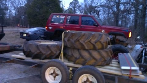 1978 Ford Bronco - A red SUV and a dark sports car are parked on a trailer loaded with large tractor tires and a motorcycle.