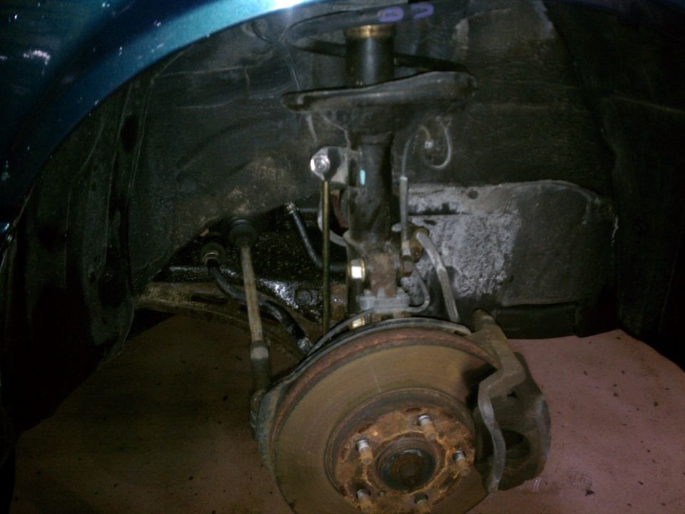 Close-up of a car's front wheel hub, brake rotor, caliper, and strut assembly. The wheel well liner is visible above