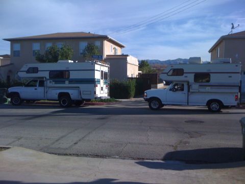 1986 Yugo GV - Two pickup trucks, each equipped with a camper shell, are parked side-by-side on a residential street.