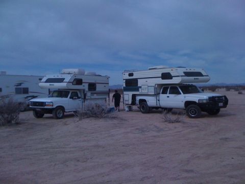 1986 Yugo GV - Two pickup trucks equipped with camper shells are parked in a sandy, desert environment under a cloudy sky.