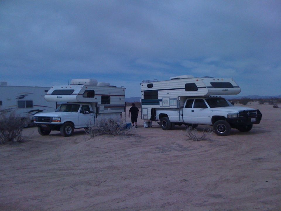 Two white pickups with slide-in campers parked on desert sand at dusk, person walking between