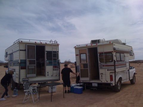 1986 Yugo GV - Two camper vans are parked in a desert setting with people interacting around them.