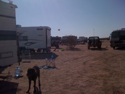 1986 Yugo GV - A desert campsite scene features multiple RVs, trailers, a dog in the foreground, and a pickup truck in the background under a clear sky.