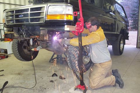1996 Ford Bronco - A man in a yellow welding jacket, gloves, and a face shield kneels beside a jacked-up dark green Ford truck/SUV, welding on the front axle/…