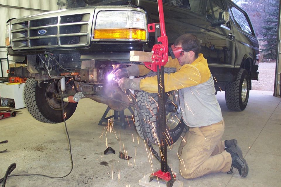 Man welding on front axle of jacked-up dark green Ford as sparks fly in a garage