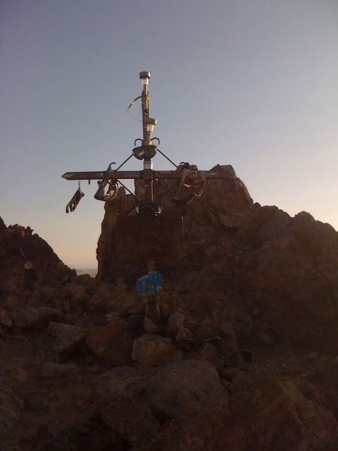 1986 Yugo GV - A summit cross stands atop a rocky mountain peak, adorned with various items and set against a clear sky at dusk.