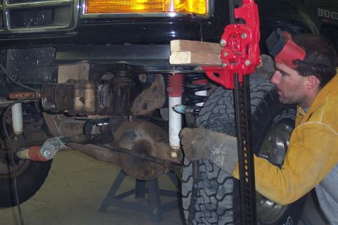 1996 Ford Bronco - Underside view of a dark-colored Ford Bronco raised on a jack stand, showing the front suspension with a new red coil spring and white shoc…