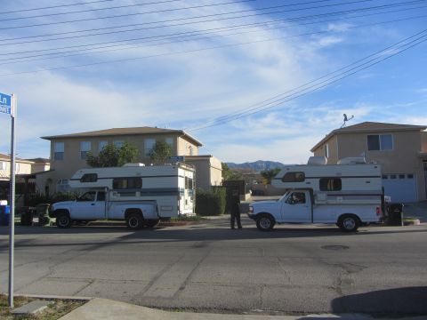 1986 Yugo GV - Two pickup trucks equipped with camper shells are parked on a street in front of houses.