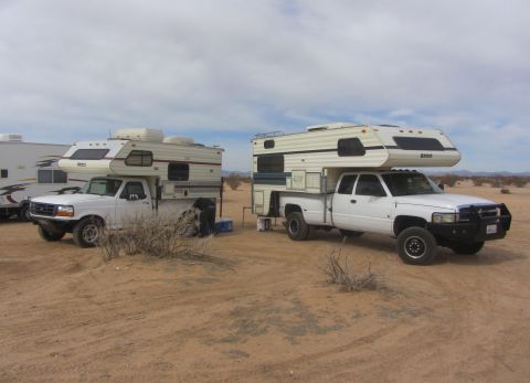1986 Yugo GV - Two pickup trucks equipped with camper shells are parked in a desert environment under a cloudy sky.