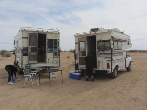 1986 Yugo GV - Two pickup trucks with camper shells are parked in a desert setting, with camping chairs and coolers nearby.