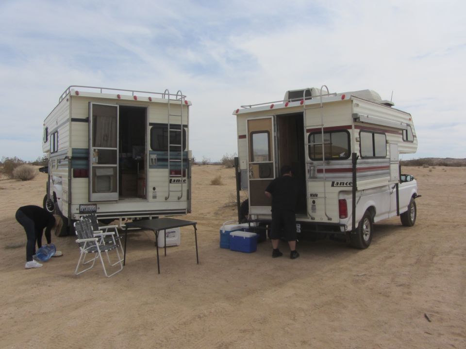 Two pickup trucks with camper shells parked in a desert landscape, with people and camping gear