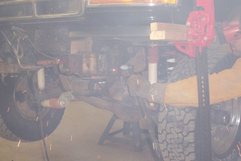 1996 Ford Bronco - Underside view of a lifted 4x4 with a person using a grinder that throws sparks near the frame, showing coil springs, red-booted shocks, of…