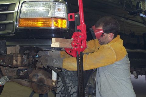 1996 Ford Bronco - A man wearing safety glasses and a tan leather welding jacket over a gray shirt uses a red Hi-Lift jack braced against a wooden block to li…