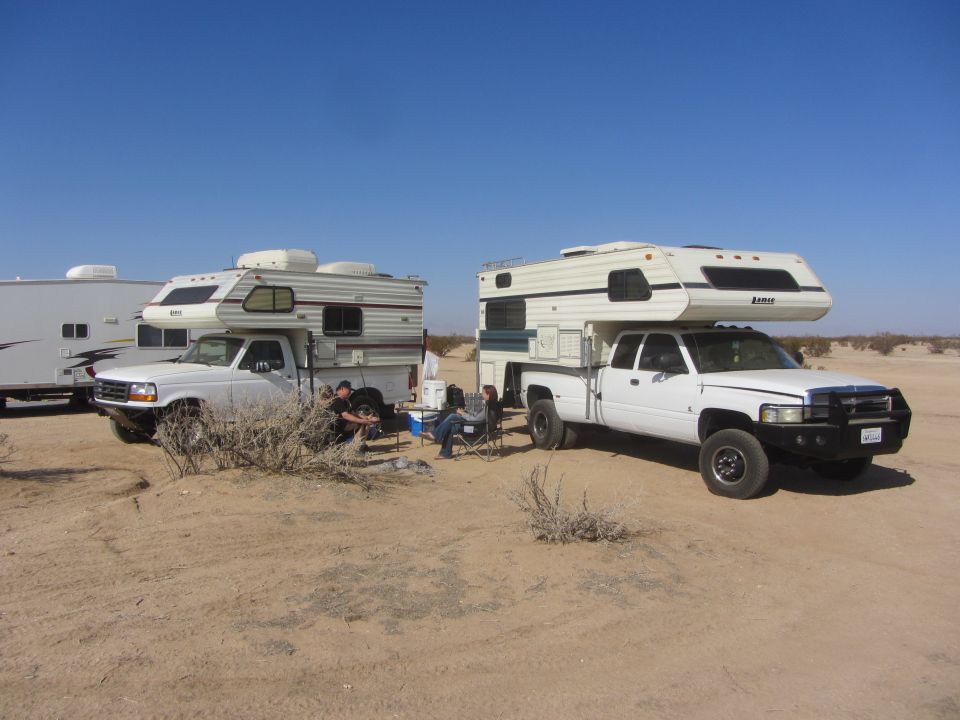 two white pickup trucks parked in a desert landscape, each with a camper shell. People are sitting in chairs near the trucks