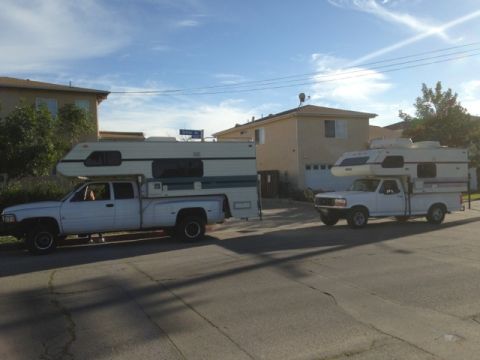 1986 Yugo GV - Two white pickup trucks, each fitted with a camper shell, are parked on a street in front of houses.