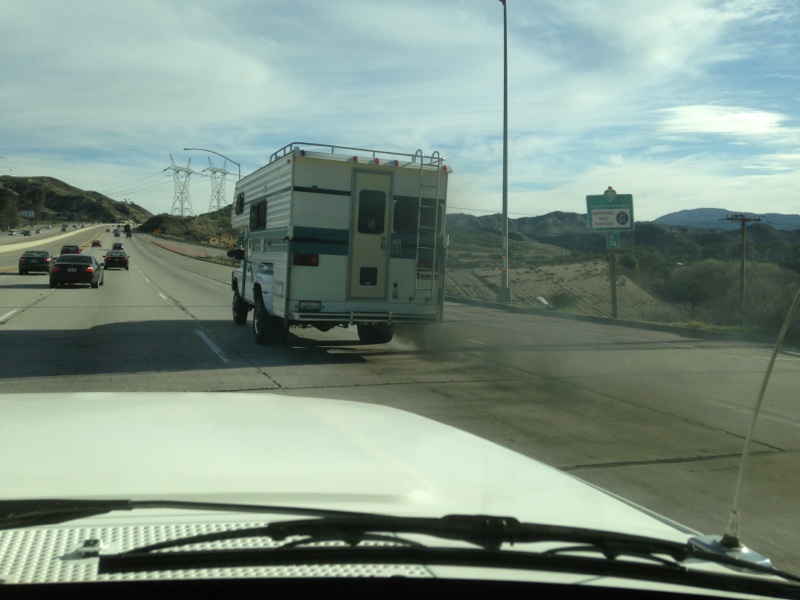 Truck camper driving on highway, seen from another vehicle. Other cars on road, power lines in background