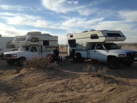 1986 Yugo GV - Two pickup trucks equipped with camper shells are parked in a desert area, with a person standing between them.