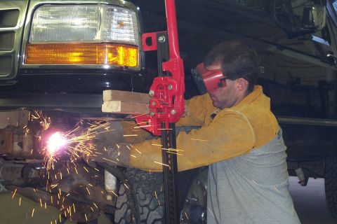 1996 Ford Bronco - A man in a yellow welding jacket and dark face shield uses a cutting or welding tool near the front bumper area of a black Ford SUV, sendin…