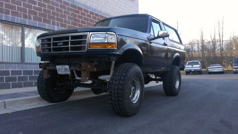 1995 Ford Bronco - This lifted Ford Bronco sits parked in front of a brick building, with other vehicles in the background.