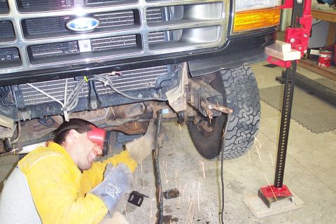 1996 Ford Bronco - A man in a yellow welding jacket, gloves, and red face shield lies on the garage floor welding or cutting under the front end of a black Fo…
