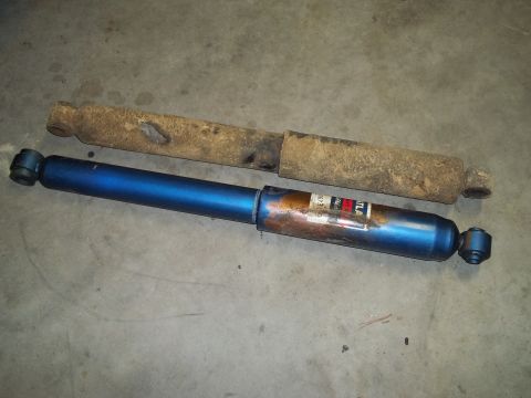 1990 Ford Bronco - Two shock absorbers, one old and rusted, the other new and blue, are laid out on a concrete surface.