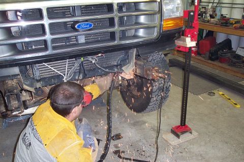 1996 Ford Bronco - A person in yellow welding gear kneels beneath the front of a black Ford truck, using an angle grinder that throws orange sparks near the f…