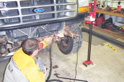 1996 Ford Bronco - A person in a yellow welding jacket kneels and uses an angle grinder, throwing sparks, while working on the front axle/steering area of a F…
