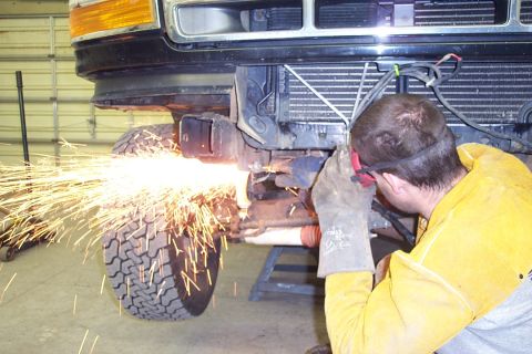 1996 Ford Bronco - A person in a tan welding jacket and heavy leather gloves uses an angle grinder or cutting tool on the front underside frame of a dark-colo…