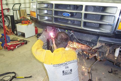 1996 Ford Bronco - A person in a yellow welding jacket and gray Fox Sports Net t-shirt welds on the front frame area of a Ford truck, with bright sparks and w…