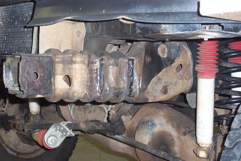 1996 Ford Bronco - Underside view of a Ford truck's frame and rear suspension, showing a rusty leaf spring shackle mount, welded frame section, and a white sh…