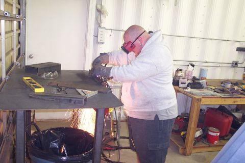 1996 Ford Bronco - A person wearing red-tinted safety glasses, a gray hooded sweatshirt, and work gloves grinds or welds metal on a steel workbench, sending a…